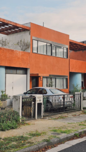 Modern orange house with aluminum fence and gate, featuring a vintage car parked in front.